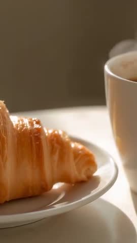Vertical panning video revealing flaky croissant on white plate with steaming coffee mug