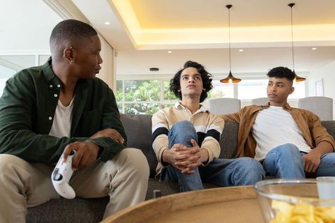 Diverse teens sitting on sofa playing video games and relaxing in modern living room