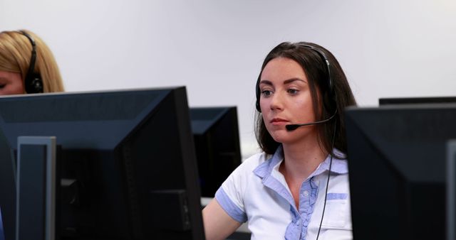 A young Caucasian woman is focused on her work at a call center, wearing a headset and looking at a computer screen, with copy space. Her profession as a customer service representative is evident from her office environment and equipment.