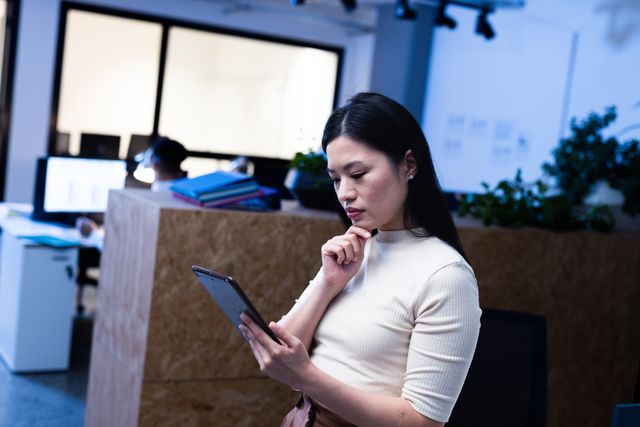 Businesswoman using a digital tablet while standing in an office at night. Ideal for illustrating concepts of working late, technology in business, professional concentration, and modern corporate environments. Suitable for articles, blogs, and advertisements related to business, productivity, and technology.