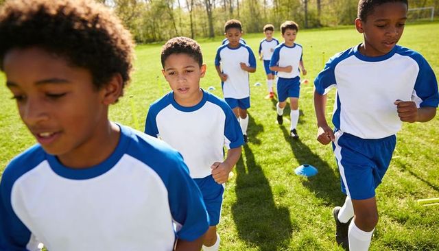 Youth soccer players running training drill on sunlit grass field with cones and poles