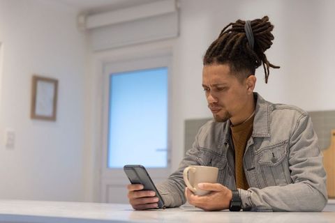 Mid-adult African American man checking phone while holding mug at bright kitchen counter