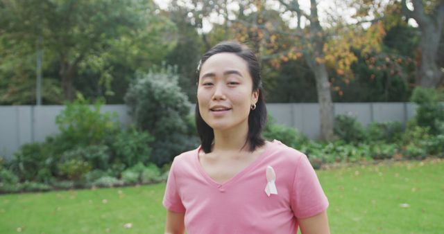 Asian woman wearing a pink shirt with a white ribbon, standing in a garden with trees and green foliage. Strong advocacy visual for breast cancer awareness, suitable for health campaigns, wellness blogs, or educational content on cancer support.