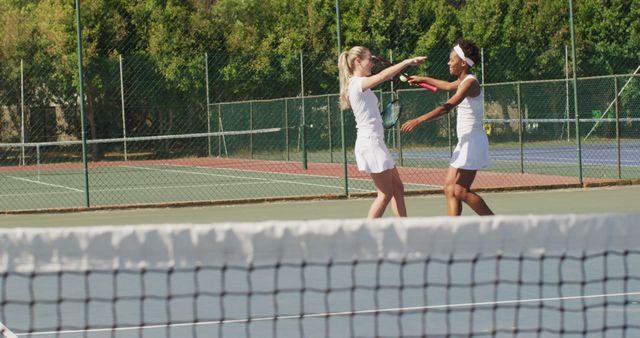 Two female tennis players in white sportswear, celebrating a victory on the court. Ideal for use in sports magazines, advertisements promoting active lifestyles, teamwork, and athletic accomplishments.