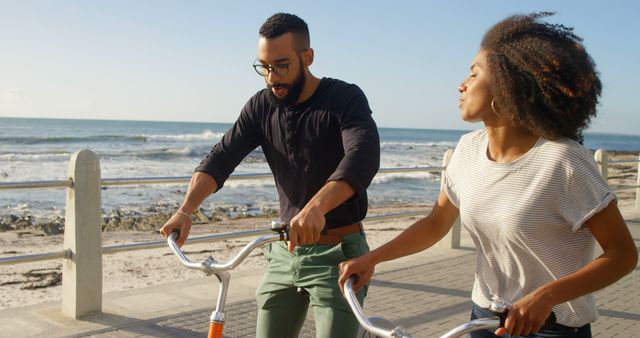 Young couple riding bicycles along a beachfront path. They are talking and enjoying the seaside view during a sunny day. Ideal for concepts of healthy lifestyle, outdoor fun, romance, and leisure activities.