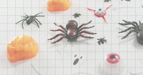 Glossy black and red toy spider sitting on white grid mat with pumpkins and eyeballs