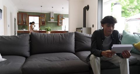 Couple relaxing at home on dark gray sofa, man reading envelope, woman working on laptop