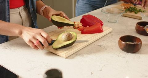 Female friends preparing avocado and bell pepper on wooden cutting board for healthy meal
