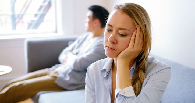 Blonde woman sitting on couch looking thoughtful with partner blurred in background. Useful for content about relationship struggles, emotional conflict, couples therapy, and personal introspection.