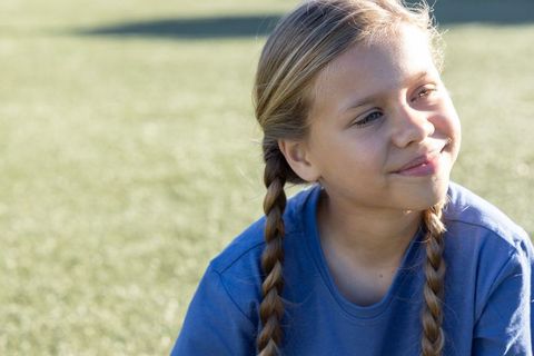 Sunlit girl with braided pigtails smiling while sitting on grassy field, outdoor childhood