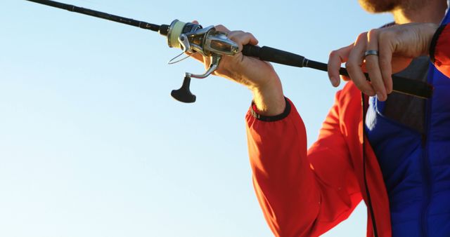 Close-up of man holding a spinning reel and fishing rod, casting outdoors. Ideal for topics related to outdoor activities, hobbies, fishing techniques, and leisure time. Can be used in advertisements for fishing gear, outdoor adventure promotions, or articles about recreational fishing.