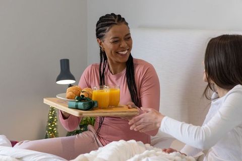 Smiling diverse female friends sharing breakfast in bed with juice, croissants and gift