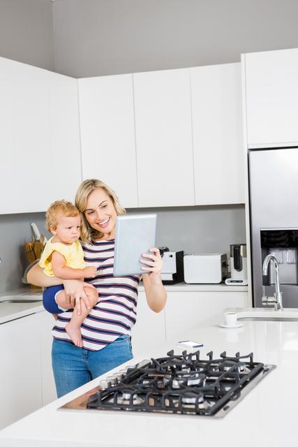 Mother holding baby girl while using digital tablet in modern kitchen. Ideal for themes related to family life, parenting, technology in daily life, and multitasking. Suitable for articles, blogs, and advertisements focusing on modern parenting, home appliances, and family bonding.