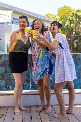 Middle-aged friends toasting yellow cocktails poolside, laughing in colorful summer cover-ups