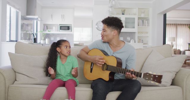 Father and young daughter enjoying music together in living room. Daughter claps while father plays guitar. Suitable for themes of family bonding, parent-child activities, home entertainment, and joyful living. Ideal for promoting music education, parenting content, and home lifestyles.