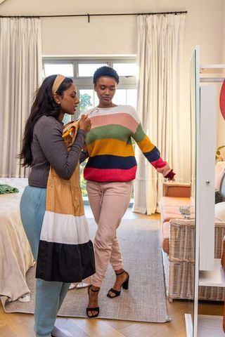 Indian and african american friends choosing dress in bedroom mirror, trying on outfit