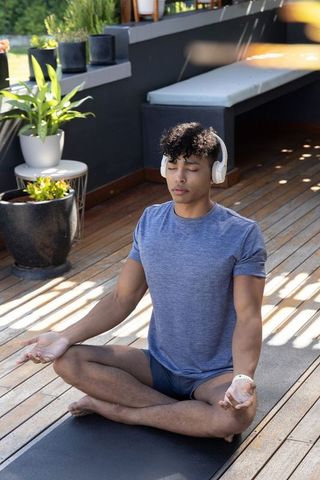 African american man meditating on wooden terrace wearing headphones on yoga mat