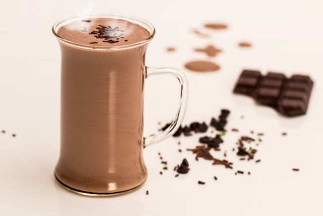 Steaming hot chocolate in a transparent mug, surrounded by chocolate pieces and sprinkled cocoa shavings on a white surface. Ideal for use in advertisements for chocolates, drink recipes, baking, desserts, or cozy winter visuals.
