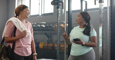 Women chatting after workout in gym featuring kettlebells, duffel and smartwatch