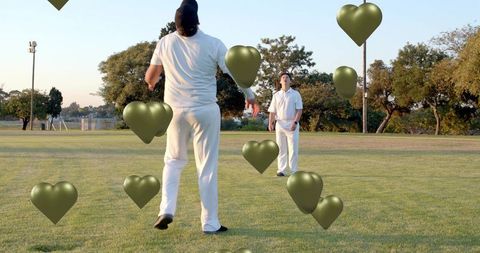 White-clad cricketers walking across turf with metallic olive heart balloons at sunset