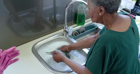 Senior woman washing tongs at soapy kitchen sink with pink gloves signaling companion care
