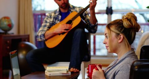 Man Playing Guitar and Woman Using Laptop in Cozy Home Environment