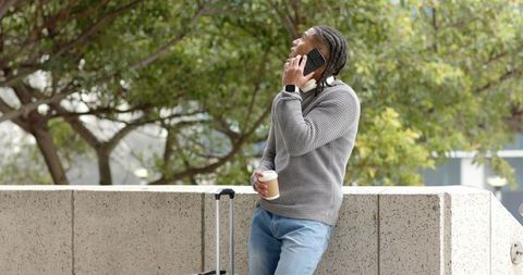 African american man leaning on urban ledge talking on phone holding coffee and luggage