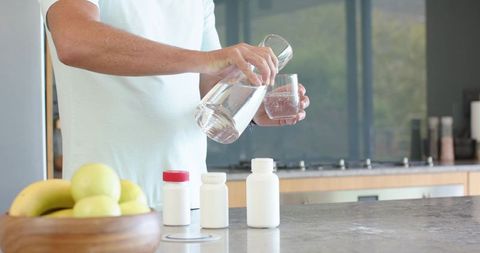 Mature man pouring water into glass near pill bottles on kitchen counter for medication