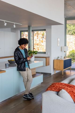 African american man leaning on pastel kitchen island using smartphone in modern open plan