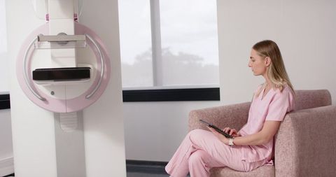 Healthcare Professional Sitting in Clinic Next to Mammography Unit Holding Tablet