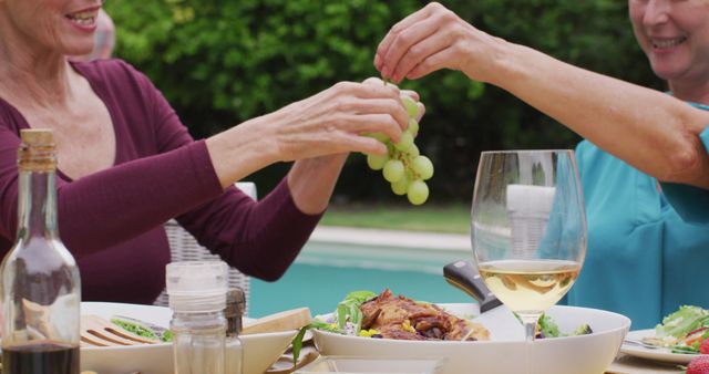 Two women are sharing grapes while sitting at a poolside table with wine, salad, and other dishes. This image can be used to promote outdoor gatherings, social events, healthy eating, or summer vibes. Perfect for advertisements, blogs, or social media posts related to food, lifestyle, or leisure activities.