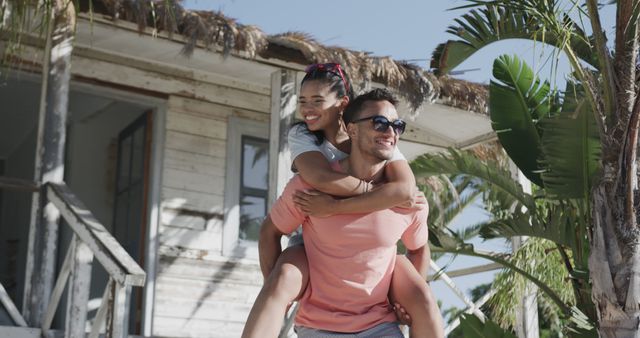 Happy couple enjoying a piggyback ride outside a vacation house with palm trees around. Perfect for travel blogs, vacation promotions, relationships, and lifestyle advertising.