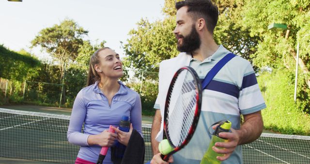 Young couple happily chatting after a friendly tennis match on outdoor court with greenery in background. They hold tennis racquets and water bottles, showcasing joy and camaraderie in sports. Ideal for use in advertisements, fitness campaigns, and social media posts documenting active lifestyles and relationships.