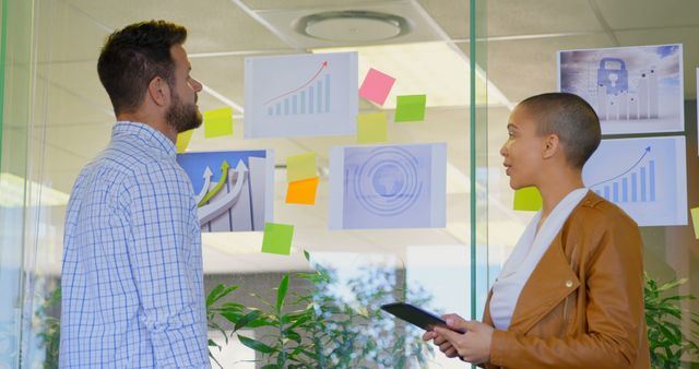 Two colleagues are discussing financial data and trends using charts and graphs on glass wall in an office meeting. They appear engaged in planning and brainstorming ideas, useful for corporate, business, teamwork, and project management related content.