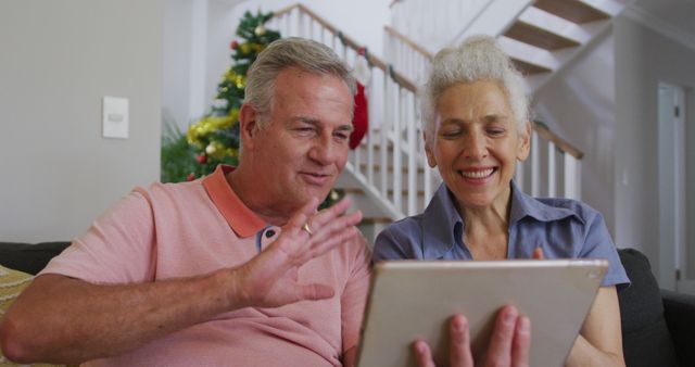 Senior couple enjoying a video call with family during Christmas using a tablet. They are sitting together at home with holiday decorations in the background. Perfect for promoting family technology use, Christmas celebrations, or senior lifestyle content.