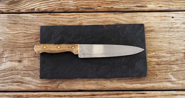 Image depicting a knife with a wooden handle and three brass rivets. The knife rests on a dark slate board placed on a wooden surface, emphasizing the rustic appeal. Ideal for use in publications or websites related to cooking, kitchenware, or rustic home decor.