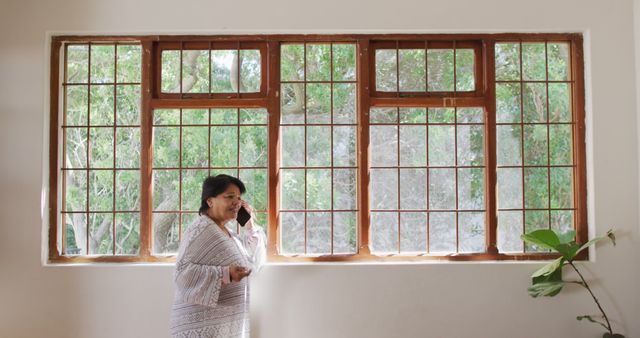 This image shows a woman engaged in a phone conversation while standing next to large windows with lush greenery visible outside. The setting is bright and airy, providing a calming and serene backdrop. Ideal for storytelling in articles or blogs about home life, communication, or relaxation. Could be used for promoting home decor, lifestyle content, or discussing the benefits of natural light in interior spaces.