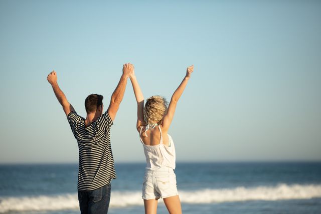 Rear view of couple standing together with arms raised, celebrating happiness at beach during sunny day. Ideal for depicting romantic relationships, vacations, summer adventures, and joyful moments. Suitable for travel websites, relationship blogs, and lifestyle advertisements.