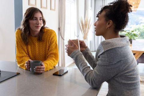 Multiracial women chatting over coffee at kitchen counter in mustard and gray sweaters