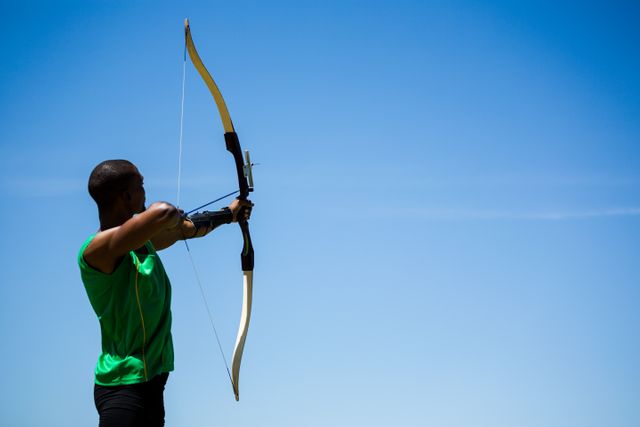 Athlete practicing archery outdoors under a clear blue sky. Ideal for use in sports training materials, fitness blogs, archery equipment advertisements, and motivational posters.