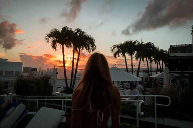 Woman with long hair enjoying the peaceful tropical sunset from a rooftop lounge surrounded by palm trees. Perfect for themes related to summer vacation, relaxation, serenity, leisure time, and tropical getaways.