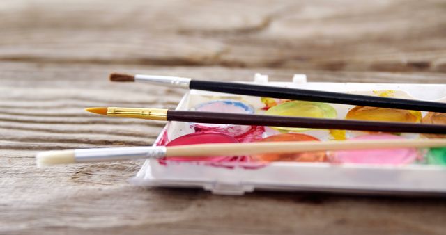 Artistic tools are laid out, highlighting paintbrushes and a watercolor palette on a rustic wooden surface. Perfect for use in articles about art supplies, creative hobbies, painting tutorials, or promoting artistic events.