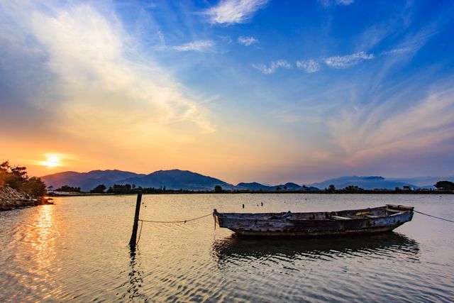 Serene lake at sunset, with vibrant sky reflecting water. Mountains in backdrop add to picturesque scene. Abandoned boat tied to old pier offers rustic charm. Ideal for travel blogs, nature inspiration, and calm ambiance design.