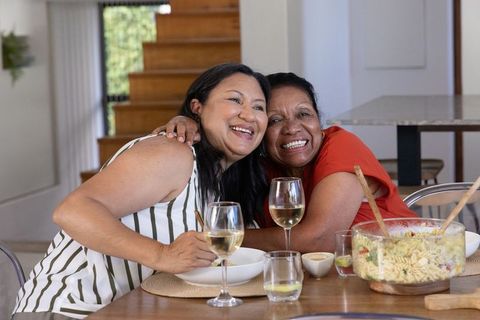 African american mother and adult daughter embracing and laughing at kitchen dining table