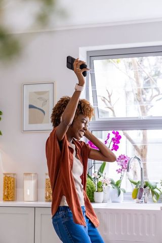 African american woman dancing and holding smartphone in bright kitchen with orchids