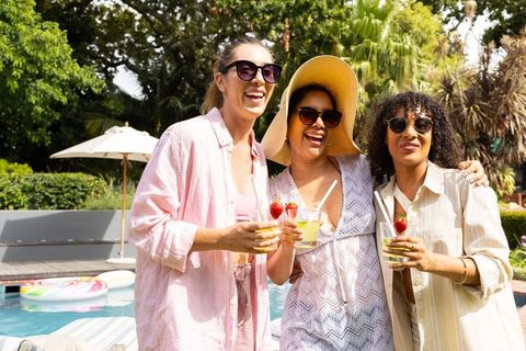 Diverse women laughing holding strawberry cocktails poolside on sunny summer vacation