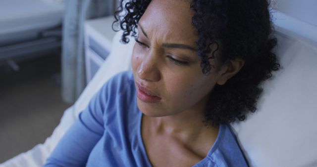 A concerned woman with curly hair is resting in a hospital bed, appearing pensive and uncomfortable. She is wearing a blue top and is in a hospital setting, showcasing a typical scene of a patient receiving healthcare. This image is ideal for use in medical articles, healthcare brochures, patient care presentations, or advertisements related to hospital services and patient recovery.