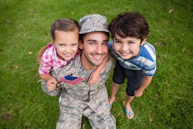 Depicts a joyful moment as a soldier reunites with his young son and daughter in a park on a sunny day. Perfect for use in themes related to military families, parental love, family reunions, or outdoor activities.