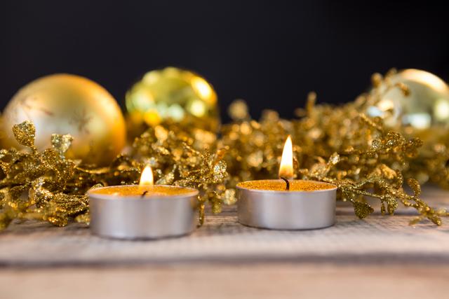 Two lit candles and golden ornaments create a warm and festive atmosphere on a wooden plank. Ideal for holiday greeting cards, festive invitations, Christmas-themed advertisements, and seasonal blog posts.