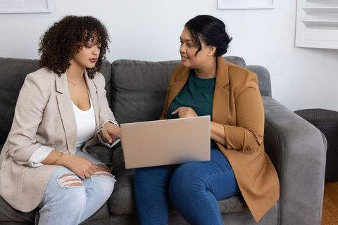 Two female colleagues collaborating on laptop while sitting on sofa in casual office